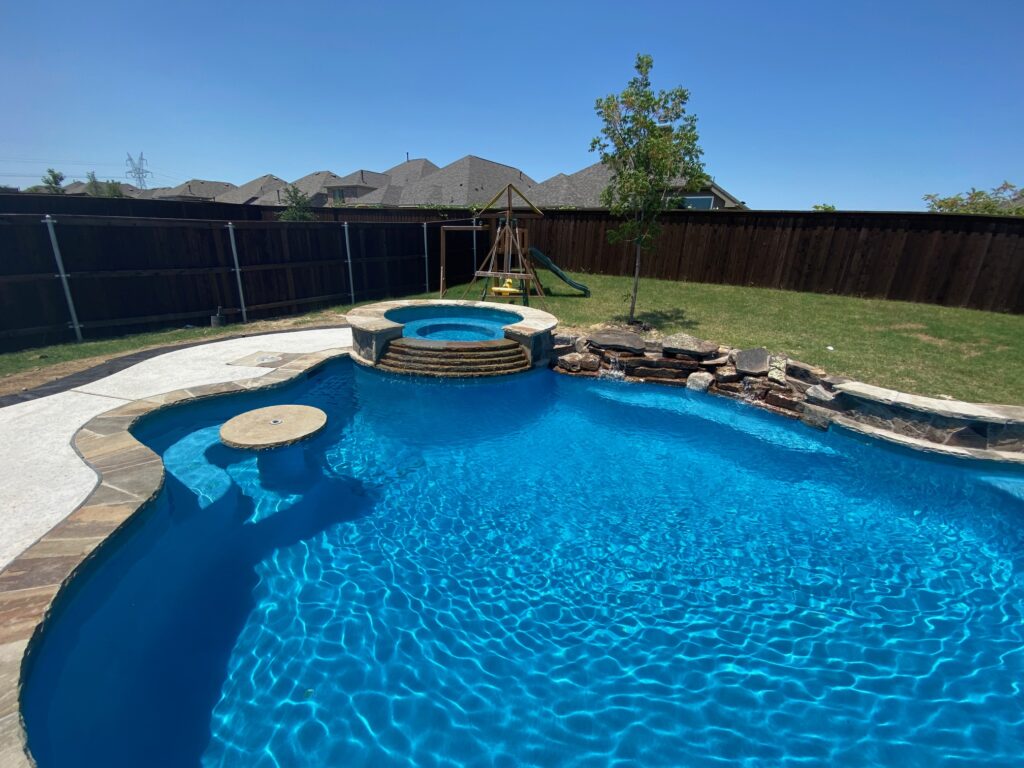 A freeform backyard swimming pool with an attached raised hot tub, a built-in swim-up table, a small rock waterfall, and a grassy lawn with a playset in the background.