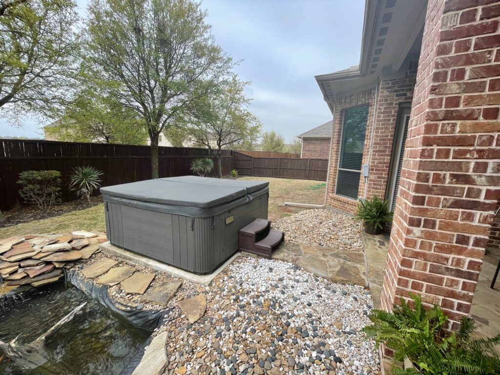 A freestanding, grey hot tub with a dark grey cover and plastic entry steps, situated in a backyard gravel landscaping bed next to a brick house and a natural pond.
