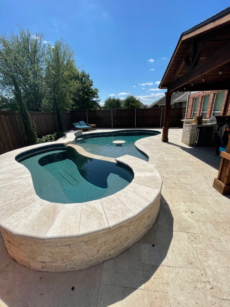 A freeform swimming pool with dark teal water, an attached raised hot tub with stone veneer, a built-in swim-up table, a large stone tile patio, and an outdoor kitchen area.