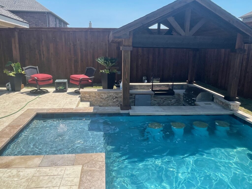 A backyard swimming pool featuring a swim-up bar with submerged stools next to a covered outdoor kitchen pavilion and lounge seating against a wooden privacy fence.