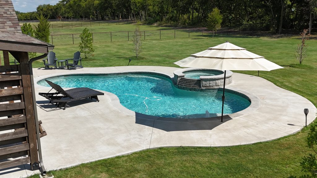 A freeform backyard swimming pool with an attached raised spa, lounge chairs, and a patio umbrella next to a large open lawn.