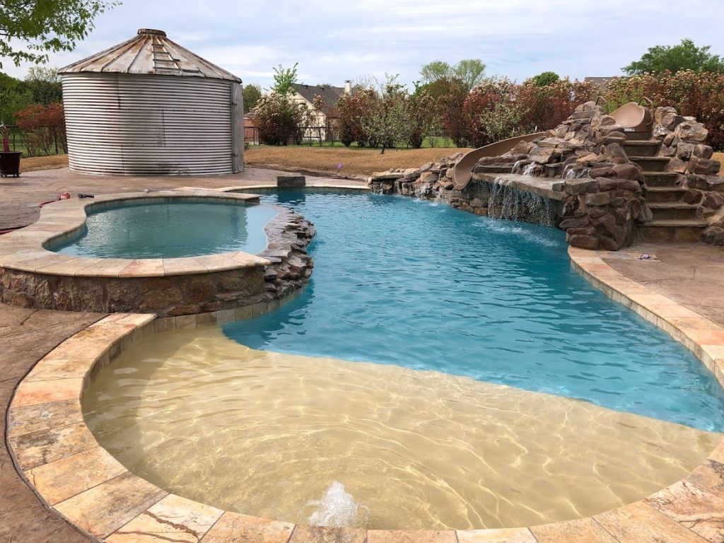 A rustic freeform swimming pool with a beach entry, a large stone waterfall with a water slide, an attached spa, and a metal grain silo in the background.